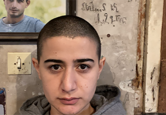 Person with short hair standing indoors against a textured wall. They wear a gray hoodie and have a neutral expression. A framed portrait of another person is visible on the wall behind them, along with light switches and decorative elements.