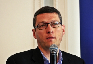 A man with short hair and glasses speaks into a microphone. He is wearing a dark blazer over a blue shirt. The background is a plain wall with a partial blue panel.