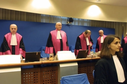 Judges in pink and black robes stand behind desks with laptops in a courtroom setting.