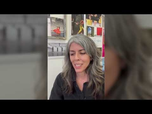 Woman with long gray hair smiling in front of a wall displaying various colorful posters.