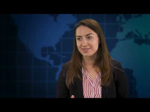 A woman in a black blazer and striped shirt speaks with a map background behind her.
