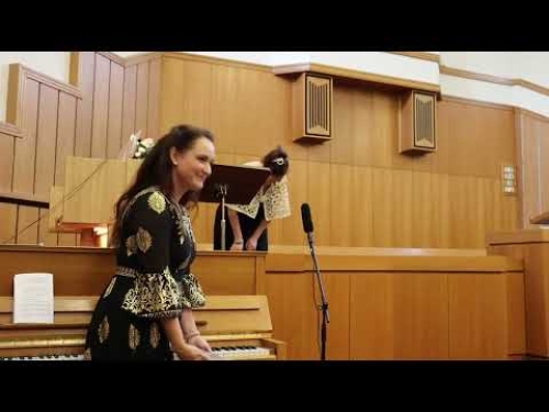 A woman in a black dress stands near a piano, with a microphone and wooden paneling behind her.