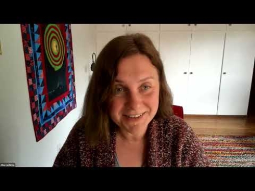 Smiling person with shoulder-length hair, wearing a patterned sweater, sits in a room with a quilt.