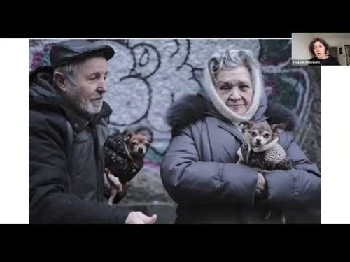 Older man and woman in coats hold small dogs; graffiti-covered wall in the background.